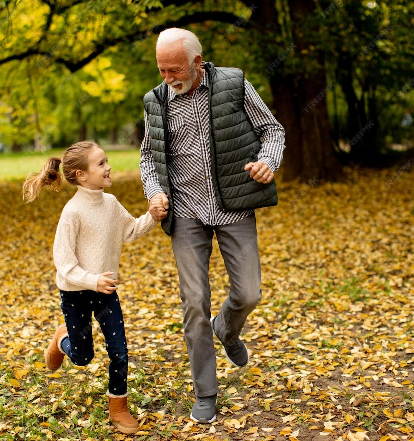 Smiling grandfather holding hands and running with young granddaughter through autumn leaves in park